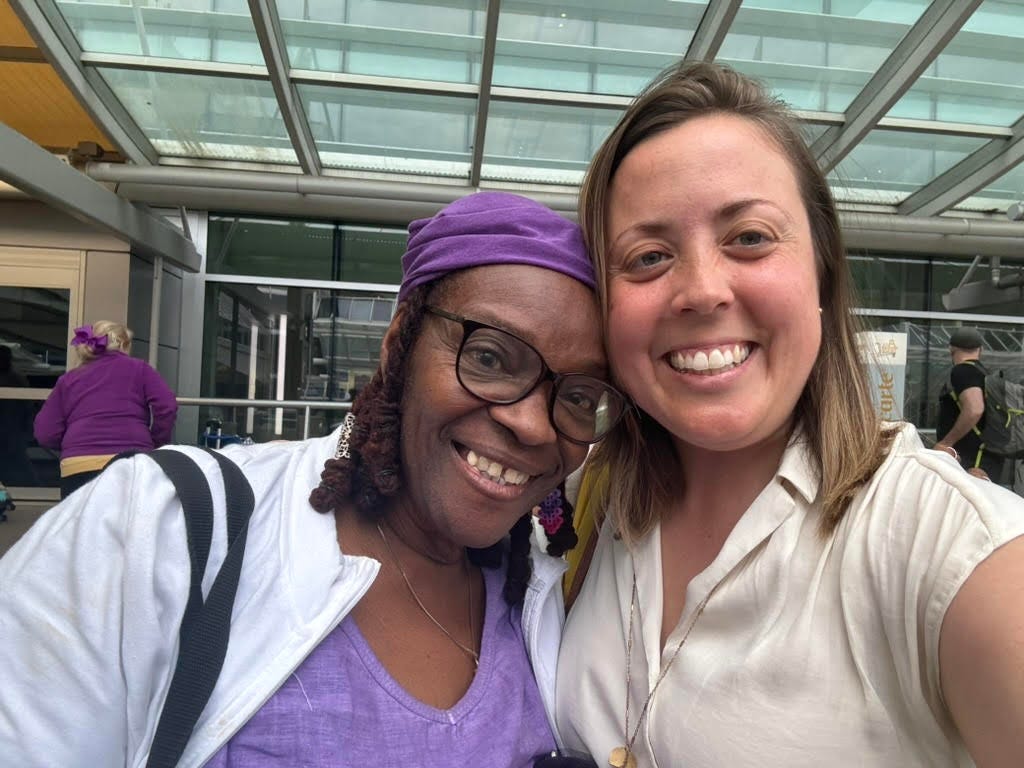 photo of Sawyer and Lillie smiling for the camera. Lillie is wearing a purple shirt and purple head cover. She has brown skin and glasses on, with a white shirt. They are both smiling big. Sawyer has shoulder length brown hair and a white blouse, with yellow, pink skin.