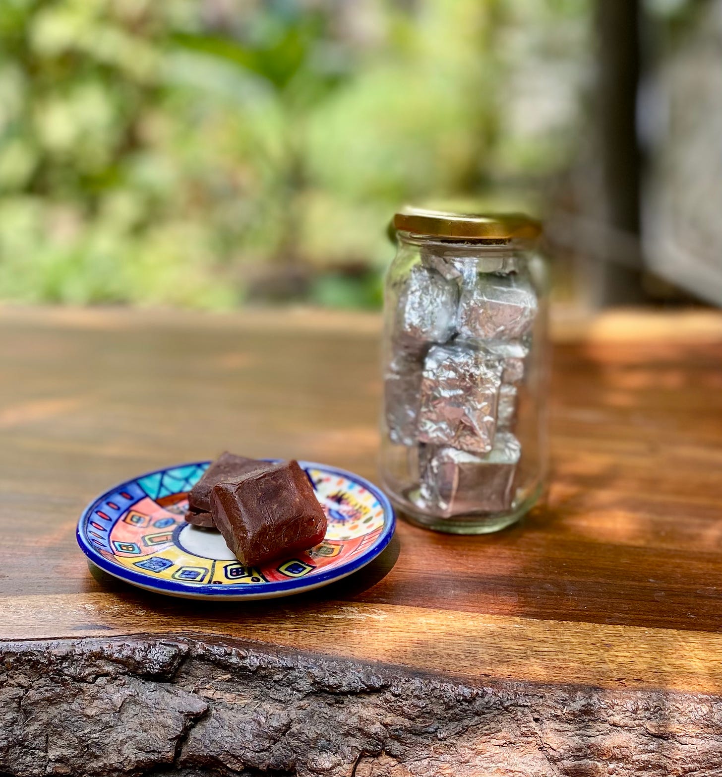 Photograph of two squares of fudge on a colourful plate and a glass bottle of foil-wrapped fudge on a rough wooden tabletop