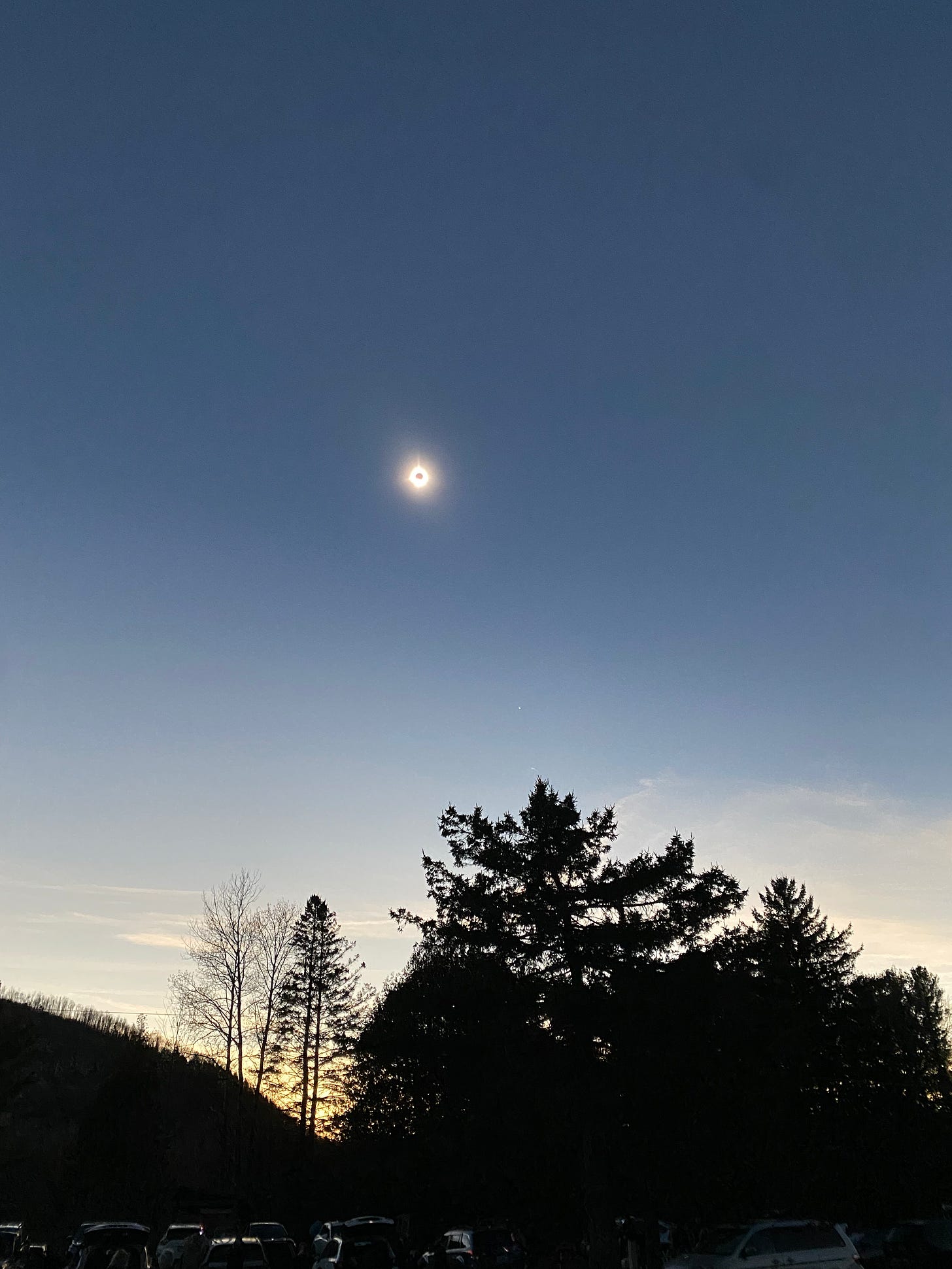 The sky at totality: the sun is a tiny gold speck with a dark dot in the middle; the sky is a deep blue with pale gold and light blue on the horizon; the trees and hillside in the foreground are silhouetted black.  
