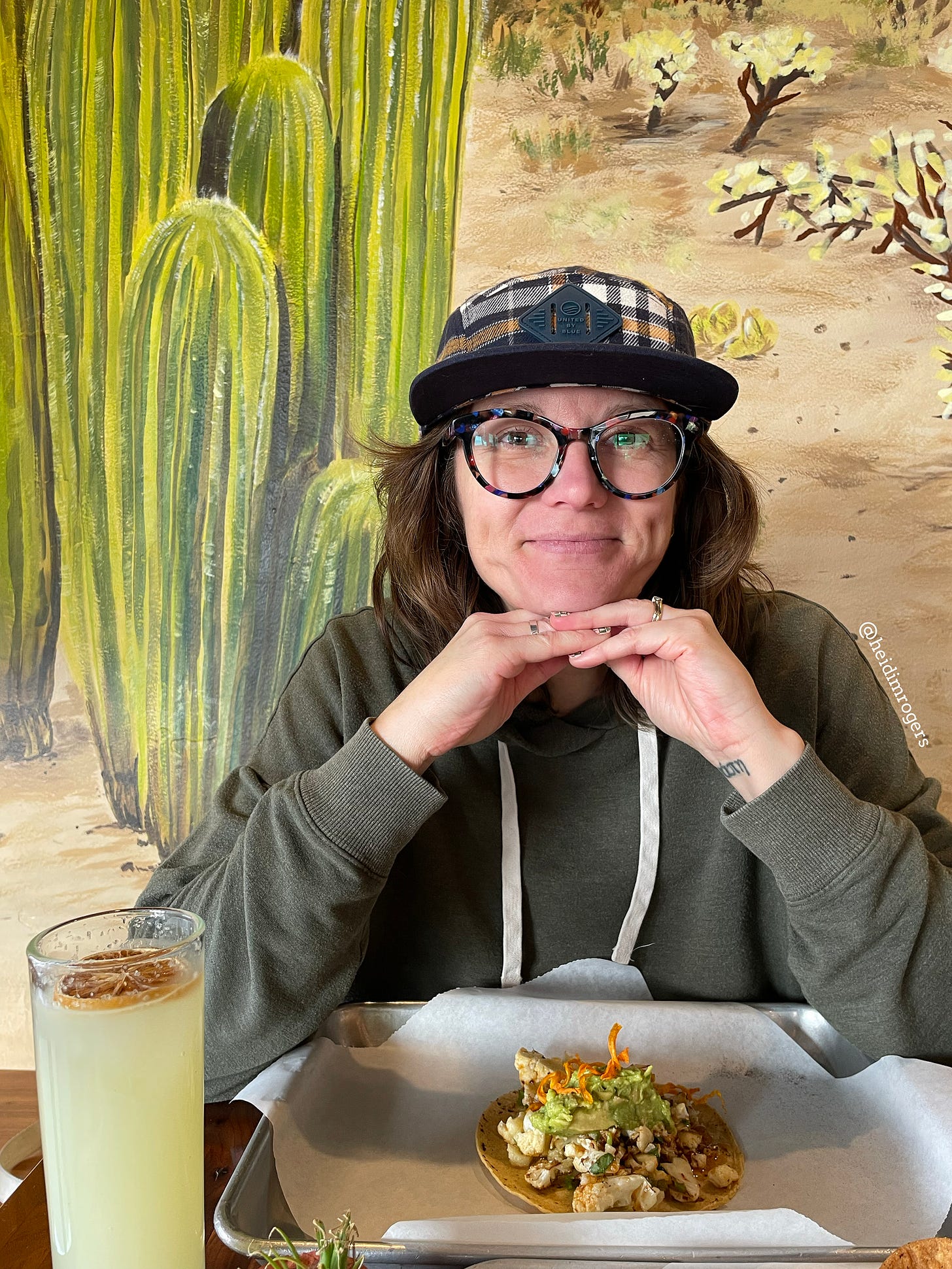 Portrait of Heidi M. Rogers, hands clasped under her chin, wearing a hat and glasses, in front of a mural with cactus and dirt. A plate of tacos and a cocktail are in front of her.