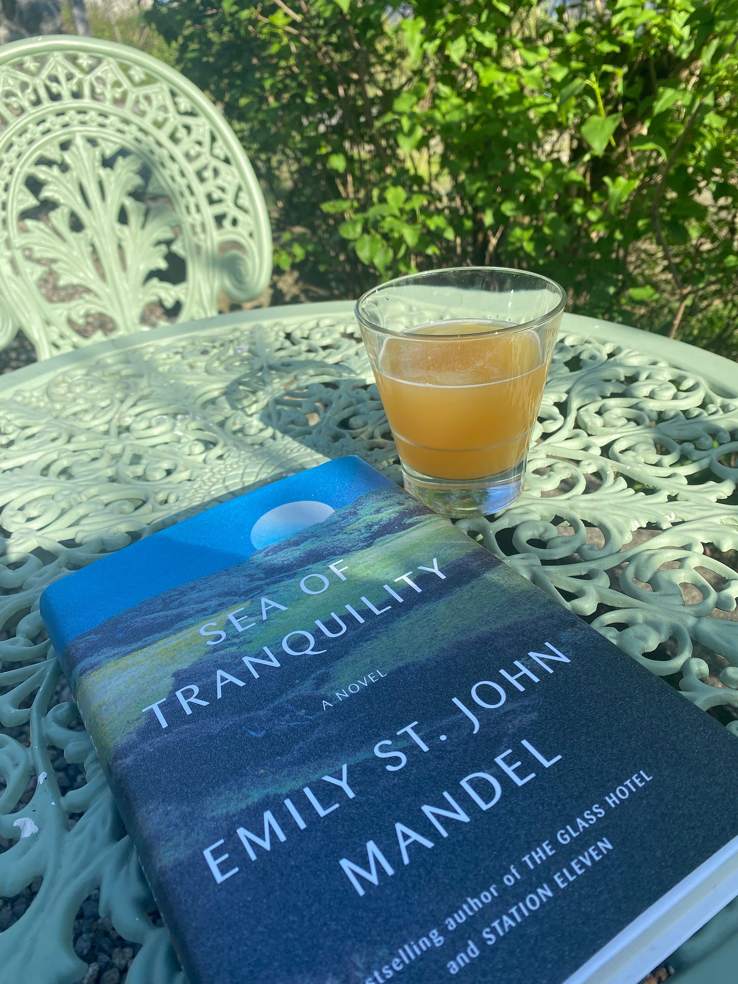 A mint green wrought-iron table in the sun. In the shaded half of the table is a glass of beer next to a hardcover novel, "Sea of Tranquility" by Emily St John Mandel. In the background is an empty chair matching the table, and leaves of a lilac bush.