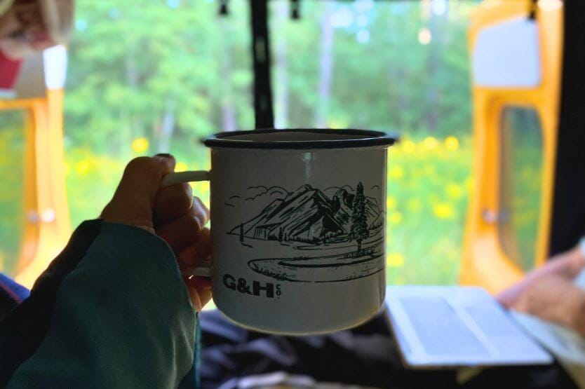 The view out of our converted camper van's back doors looking over yellow wildflowers as I hold a mug of coffee in the foreground