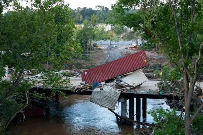 A bridge has collapsed over a river, with shipping containers and debris scattered on and around it. Trees surround the bridge, and construction equipment is in the background