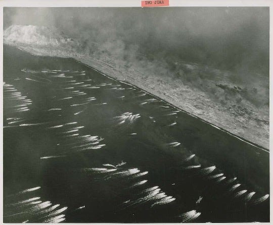 This is a historic black-and-white aerial photograph of the Battle of Iwo Jima during World War II. The image captures the moment U.S. Marines and landing craft approach the island's shoreline under heavy fire. Mount Suribachi, the island’s dominant volcanic peak, rises in the background, partially shrouded in smoke from ongoing bombardments. The dark sands of the beach contrast sharply with the white surf and the wakes left by the landing craft. Plumes of smoke rise from various points on the island, indicative of the intense combat and bombardment preceding the invasion. The photograph vividly conveys the scale and ferocity of the battle, one of the most significant and costly engagements in the Pacific theater.