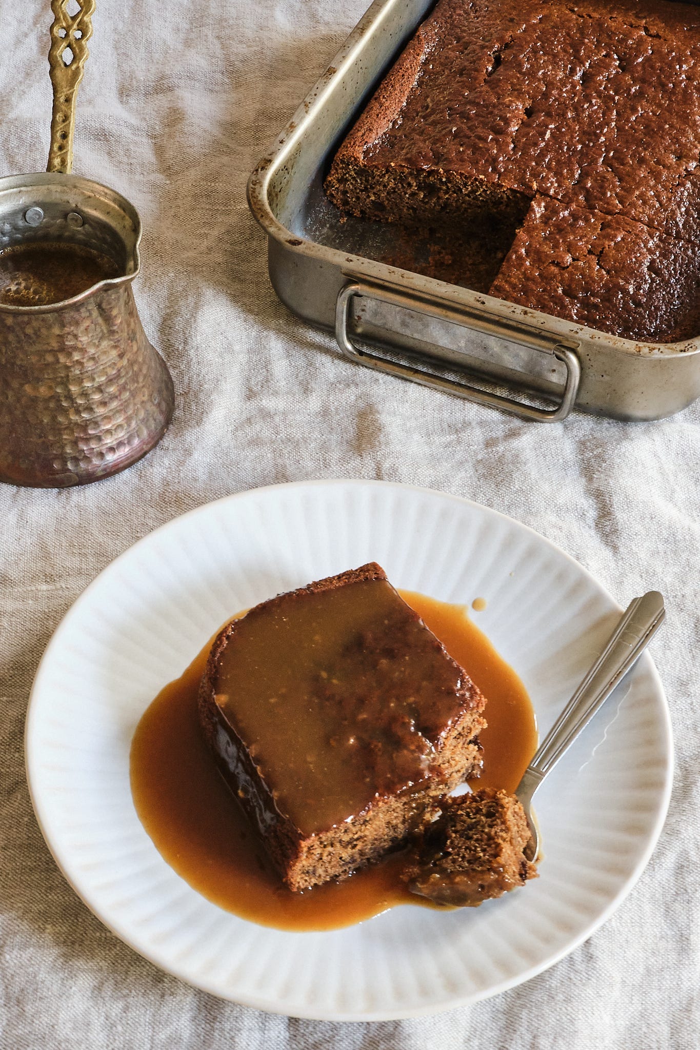 Prune & Chinese Five Spice Sticky Toffee Pudding with Miso Butterscotch Sauce baked in a tin, with a slice taken out and served on a white plate.