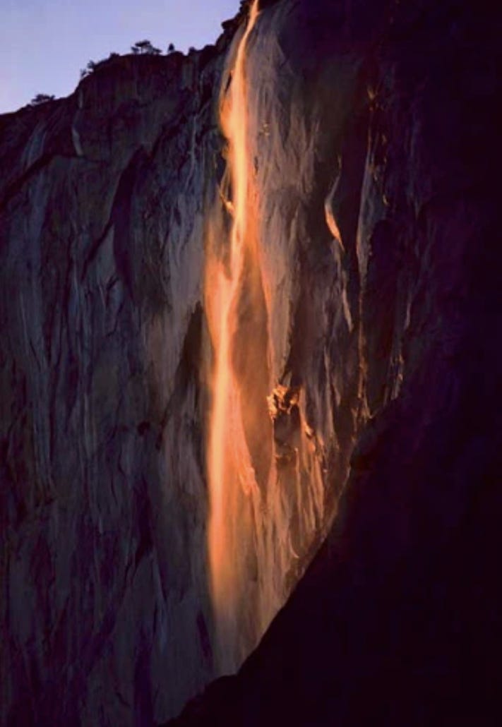 A thin waterfall cascades off a cliff wall, glowing red thanks to the sun's illumination