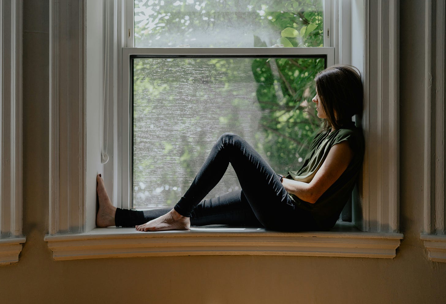 A young white woman wearing a sleeveless shirt and dark jeans sits on a window sill looking outside. 