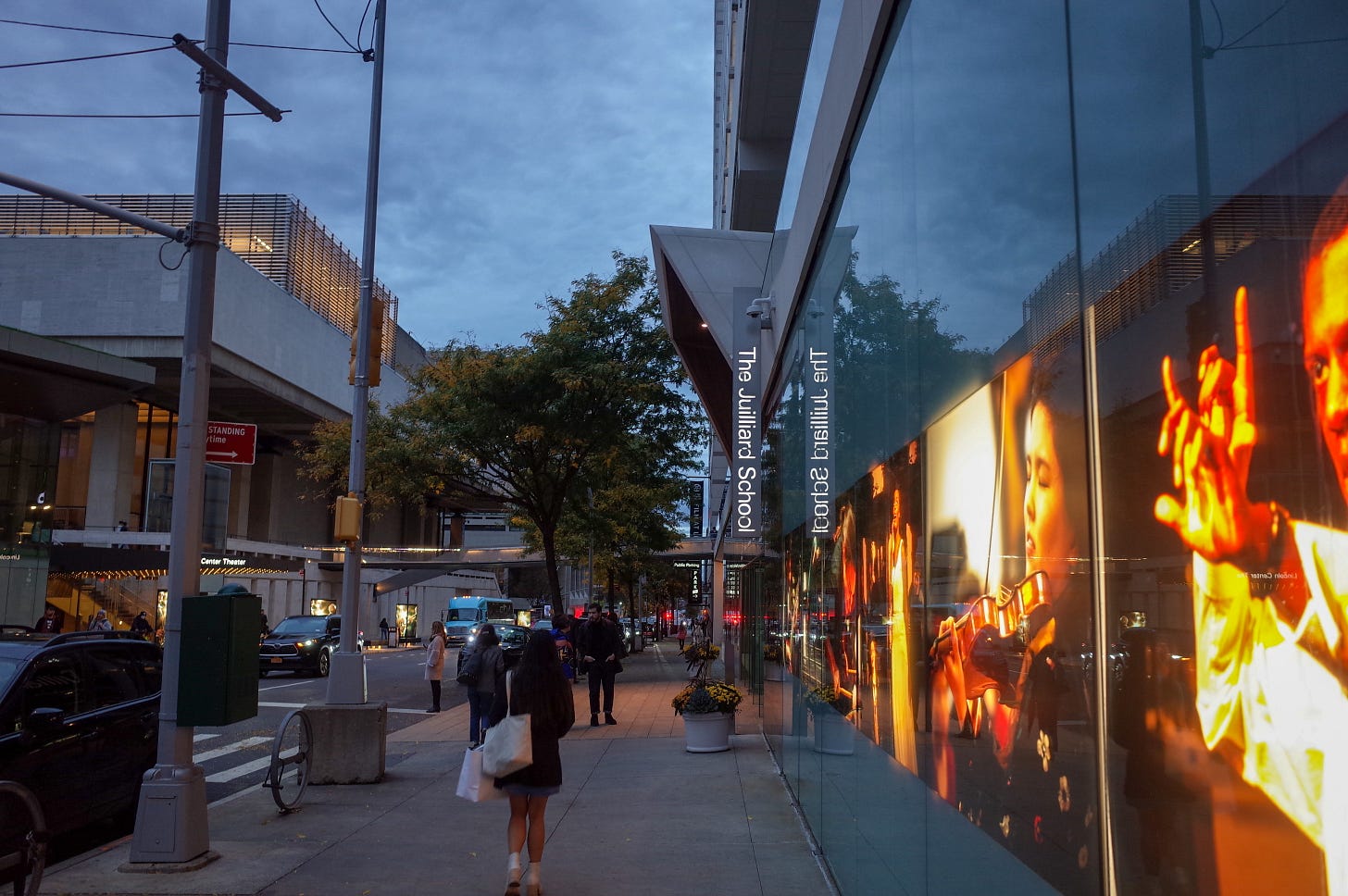 A wall lit up with large pictures of Juilliard student performers in sepia tones follows a cement sidewalk. A street with cars is visible to the left of the picture, people are on the sidewalk, and the letters of the vertical “The Juilliard School” sign are lit up near the center of the picture.