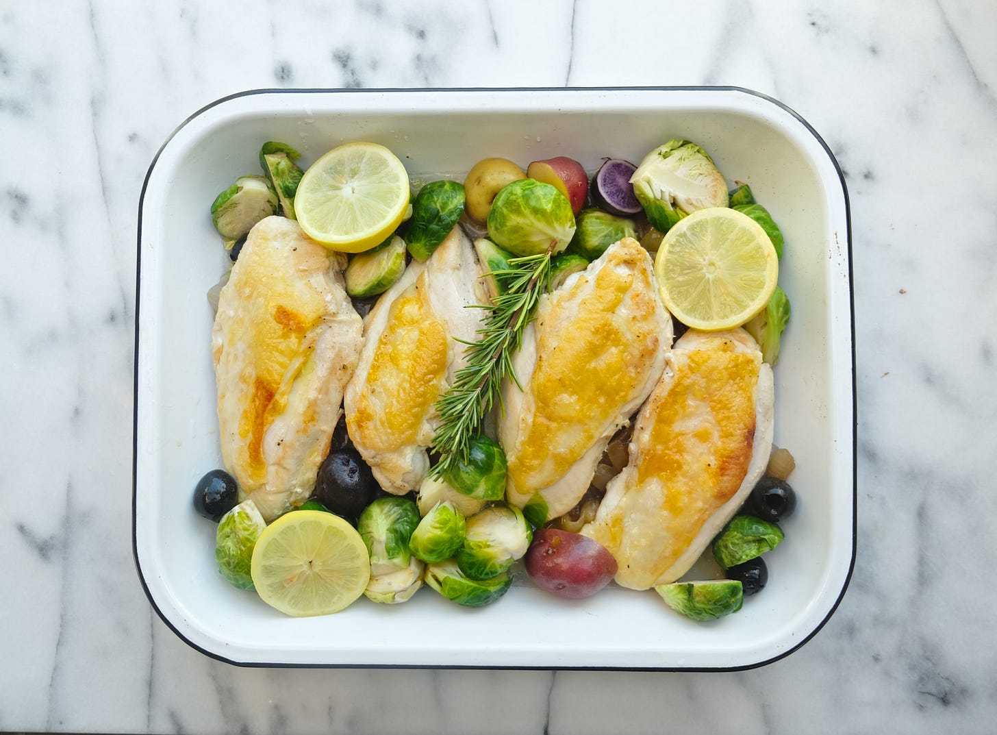 4 skin-on chicken breasts in a baking tray with roasted Brussels sprouts, fingerling potatoes, and lemon wedges