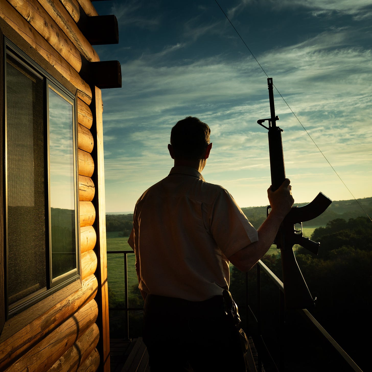 The silhouette of a man standing beside a log structure holding a gun.