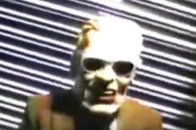 Low quality image of someone in a ‘Max Headroom’ disguise grinning for the camera, slightly skewed perspective.