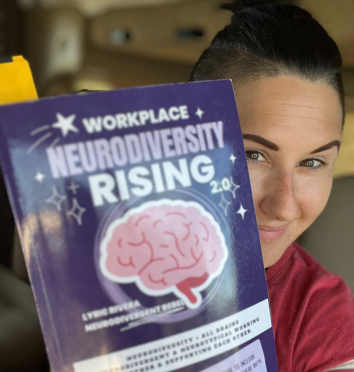Lyric Rivera, holding up a purple book with a pink brain on the cover, smiling from behind it.  