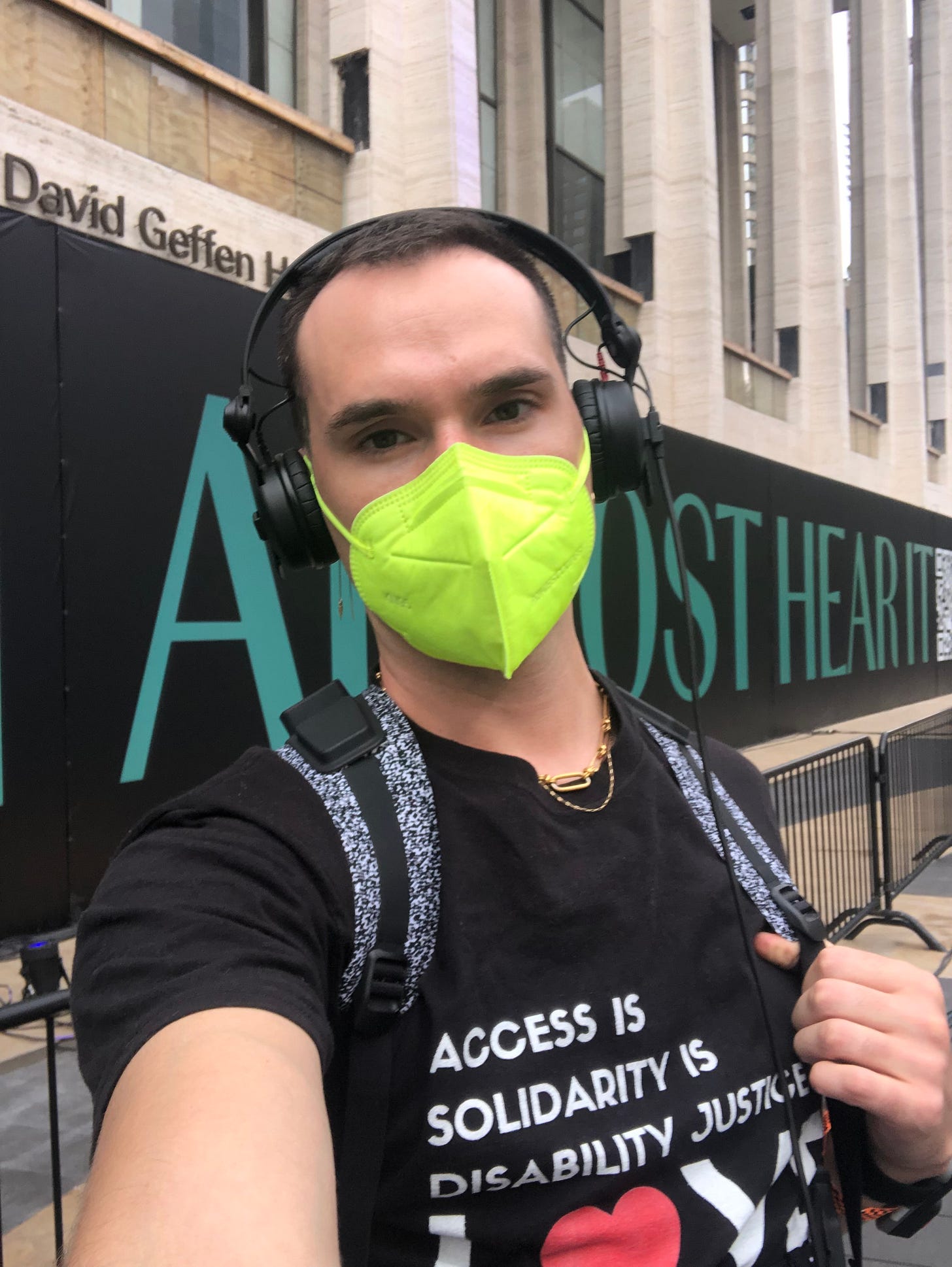 A white person is wearing over-the-ear headphones and a bright neon green KN95 mask outside at Lincoln Center. They are wearing a shirt that says Access is solidarity is disability justice and there are straps over it from a vest that pumps bass.