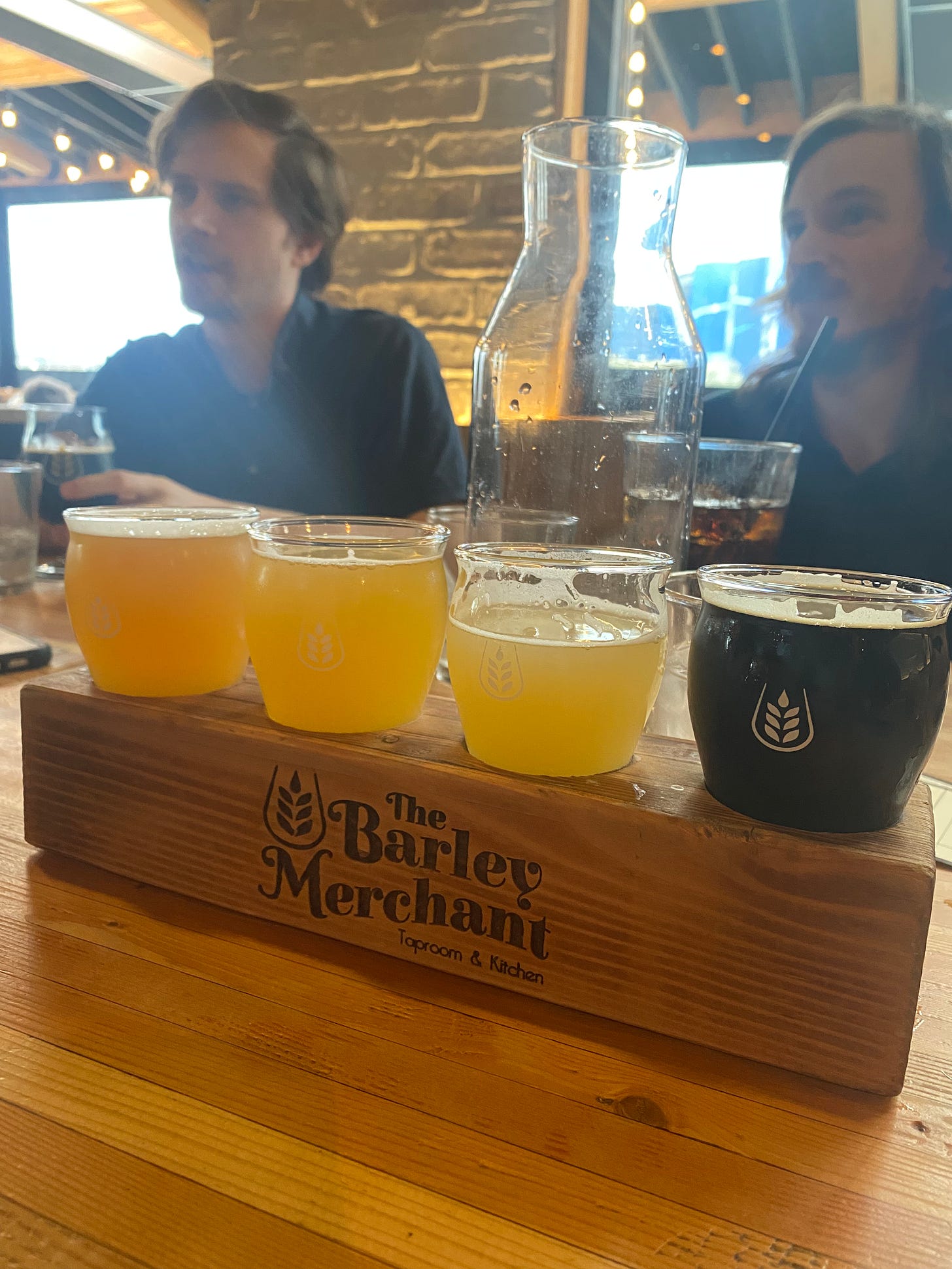 A flight of beer in a wooden block etched with the Barley Merchant's name and logo, a tulip glass with the shape of a sheaf of barley inside.  Most of the beers are light and hazy in colour, with a dark stout at the end. In the background across the table are my brothers, sitting in front of a brick wall.