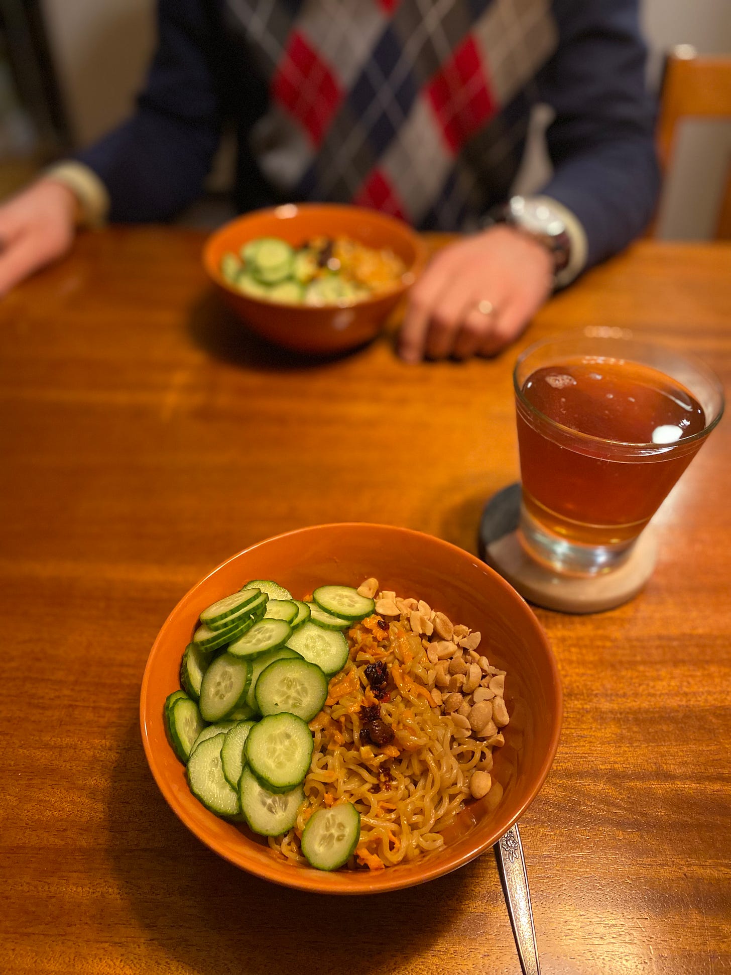 Two orange bowls of the sesame noodles described above, mixed with shredded carrot and topped with cucumber slices, peanuts, and chili crisp. On a coaster is a glass of amber ale. Jeff sits across the table, his hand resting next to his bowl.