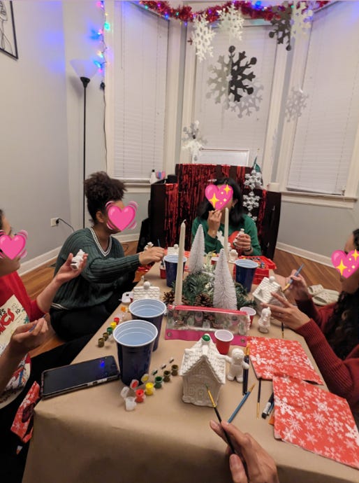Friends sit around a table filled with Christmas decorations like snowflakes hanging from the ceiling. Everyone is holding a paintbrush, getting reading to paint their white ceramic gingerbread houses