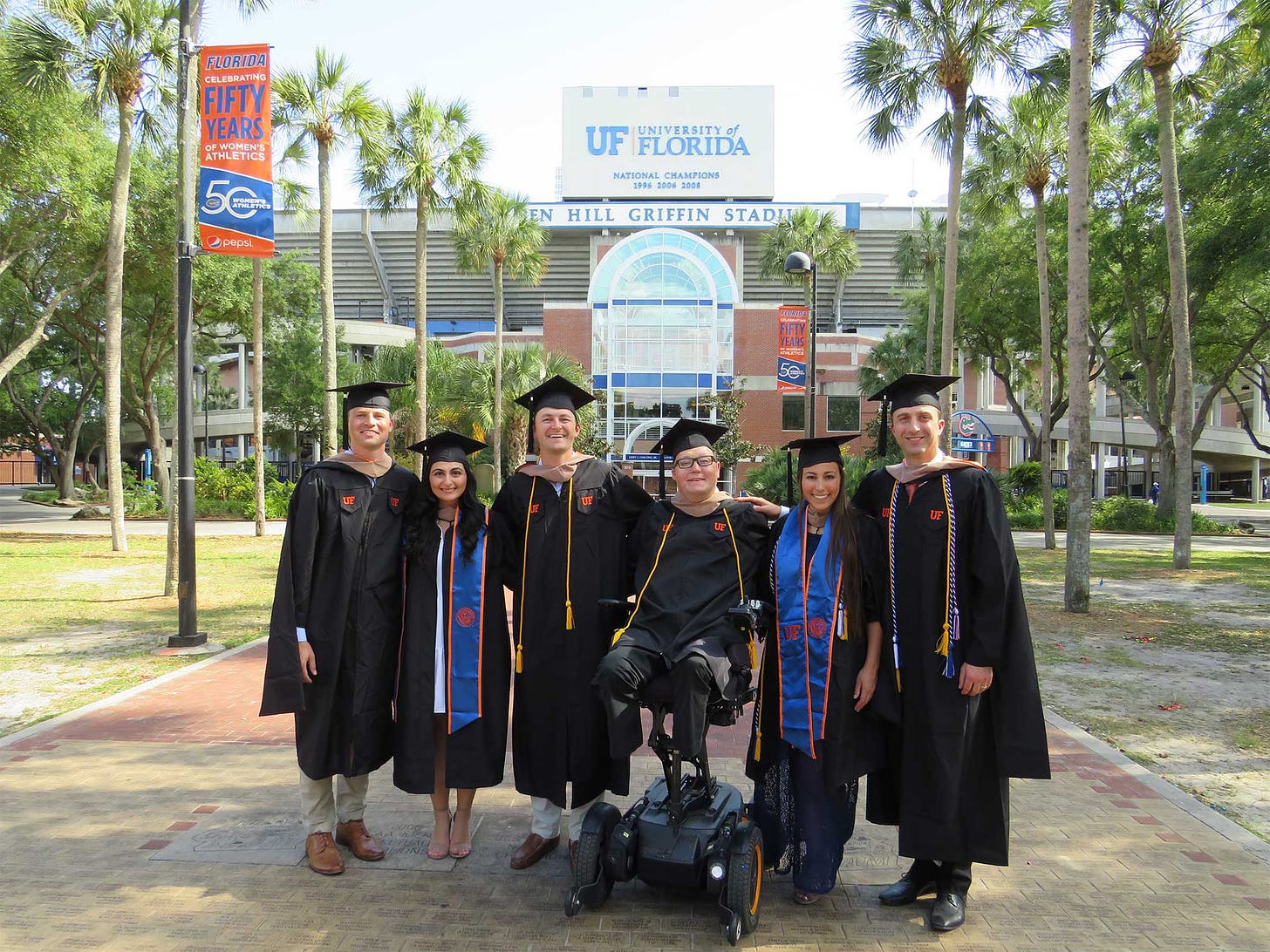 John wearing a graduation gown, pictured with fellow alumni in front of the University of Florida football stadium.