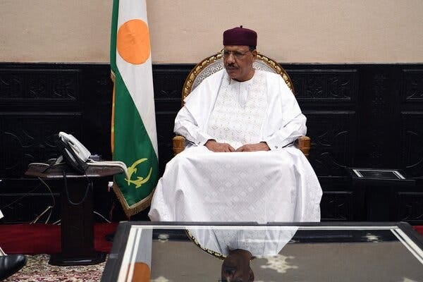 A man sits in a chair beside a flag of Niger.