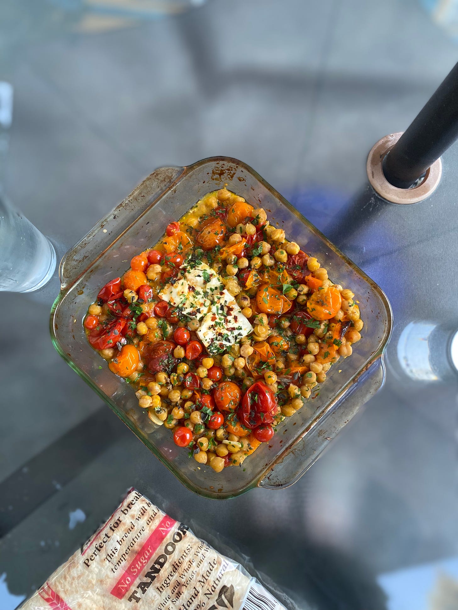 A clear, but stained, square pyrex baking dish full of orange and red small tomatoes mixed with chickpeas. In the middle is a somewhat rectangular piece of feta. Everything is sprinkled with chili flakes and chopped herbs. Just visible in the bottom of the photo is a package of taftoon.