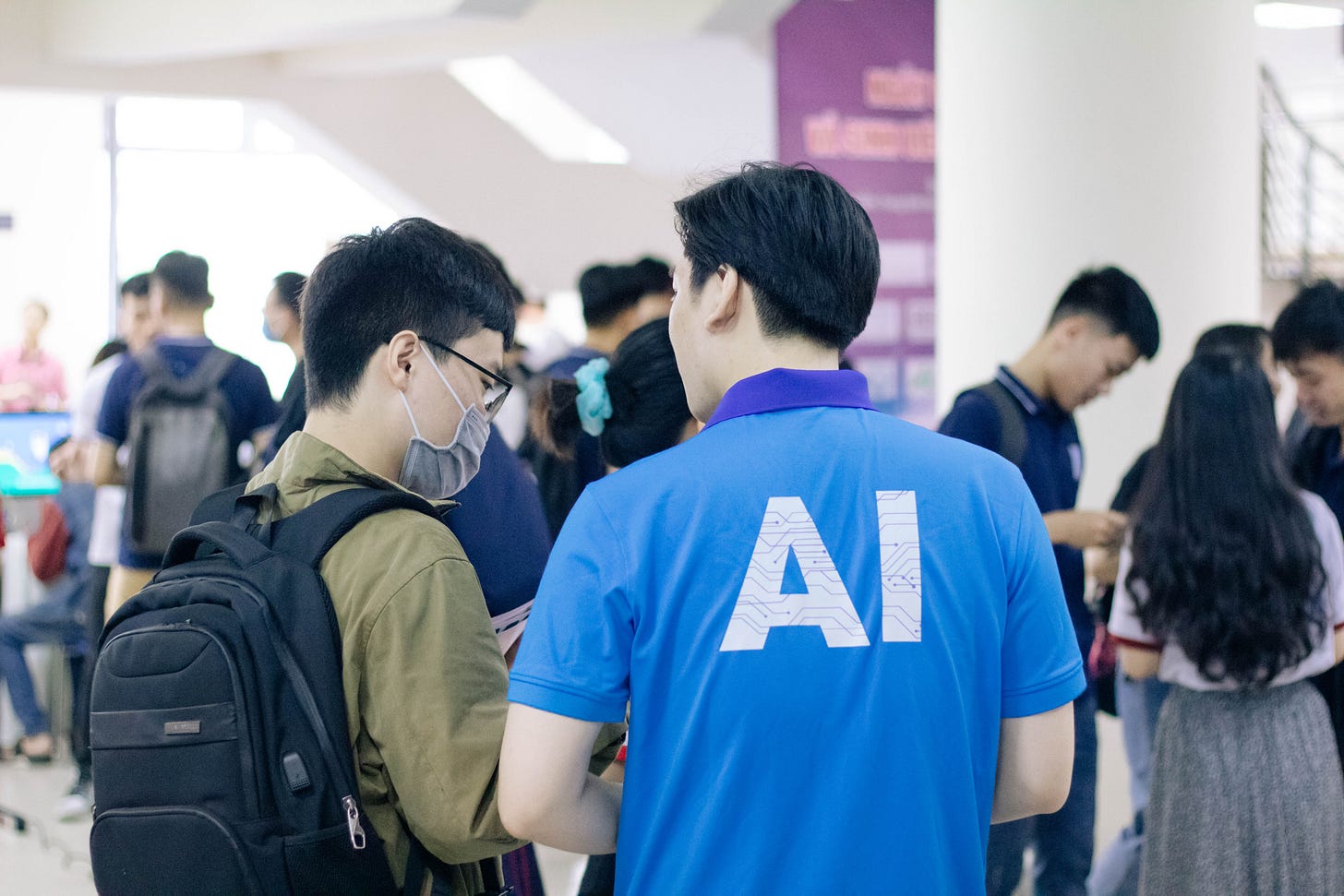 Alt-text: Foto em cores de um homem chinês, de costas, com AI escrito na camiseta azul. Ele fala com outro homem chinês, de máscara no rosto e mochila nas acostas. Ao fundo, diversos jovens conversam em grupos. Alt-text: Foto em cores de um homem chinês, de costas, com AI escrito na camiseta azul. Ele fala com outro homem chinês, de máscara no rosto e mochila nas acostas. Ao fundo, diversos jovens conversam em grupos.
