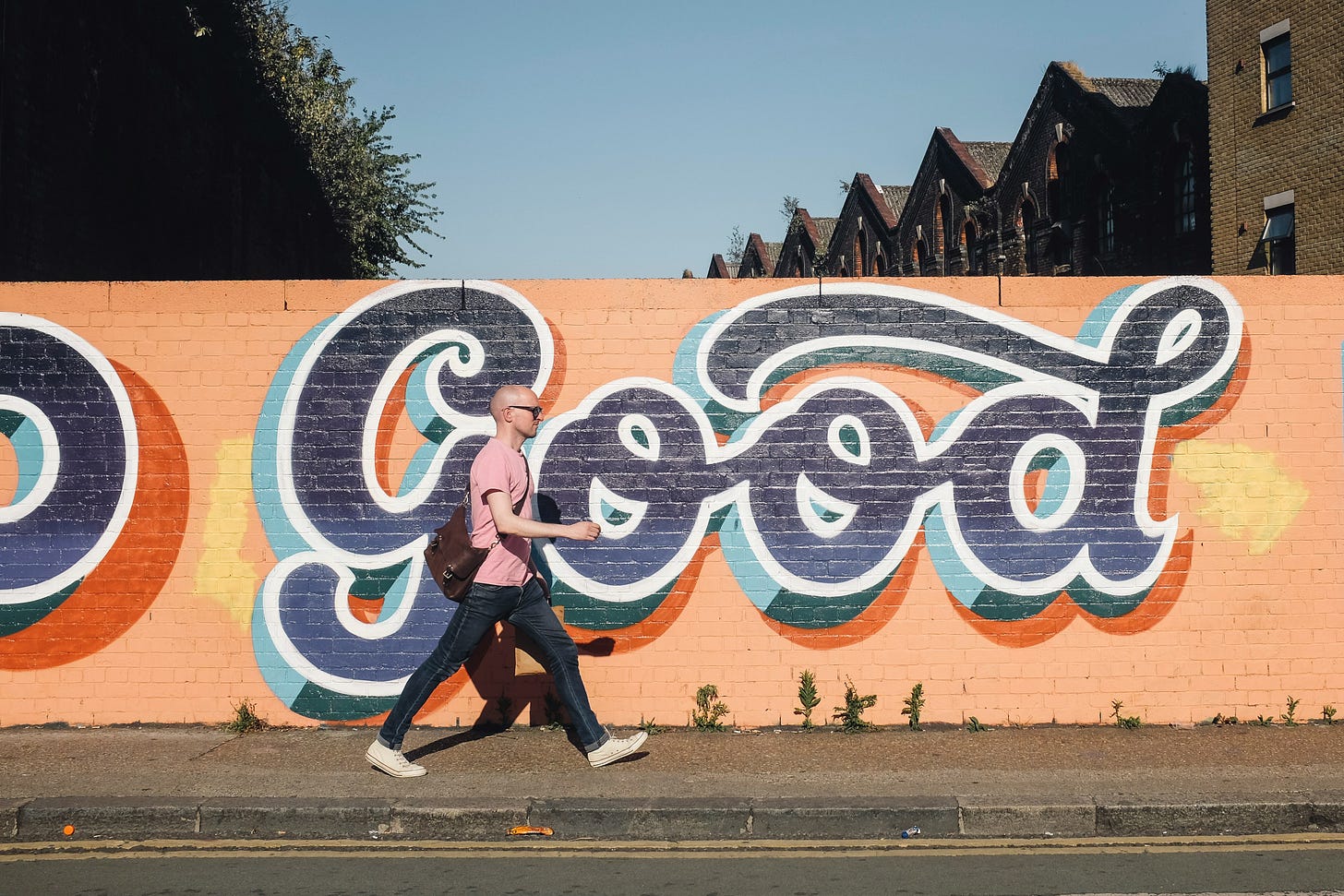 A person wearing a pink shirt, jeans, sunglasses, and a shoulder bag walking in front of an orange wall with the word "good" written on it in blue block letters