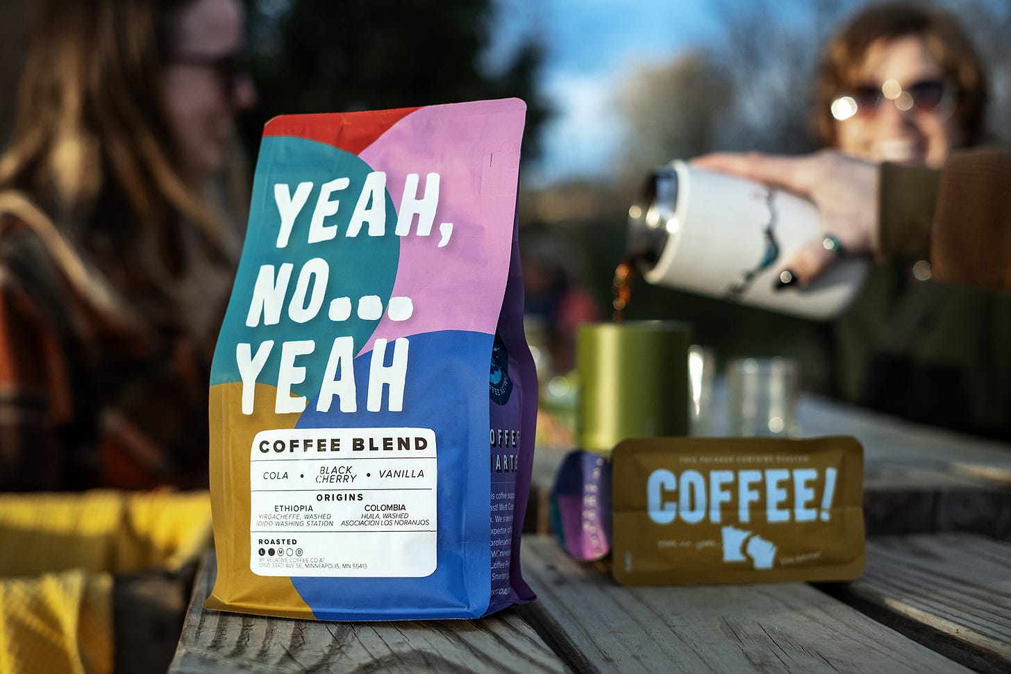 A close up of a brightly colored bag of coffee with the title Yeah, No...Yeah Coffee on a wooden picnic table. A second bag is turned on its side with the bottom facing out designed with the word "Coffee!" and the states of WI and MN in white silhouette. In the background, friends pour coffee into a mug and smile but are blurred.