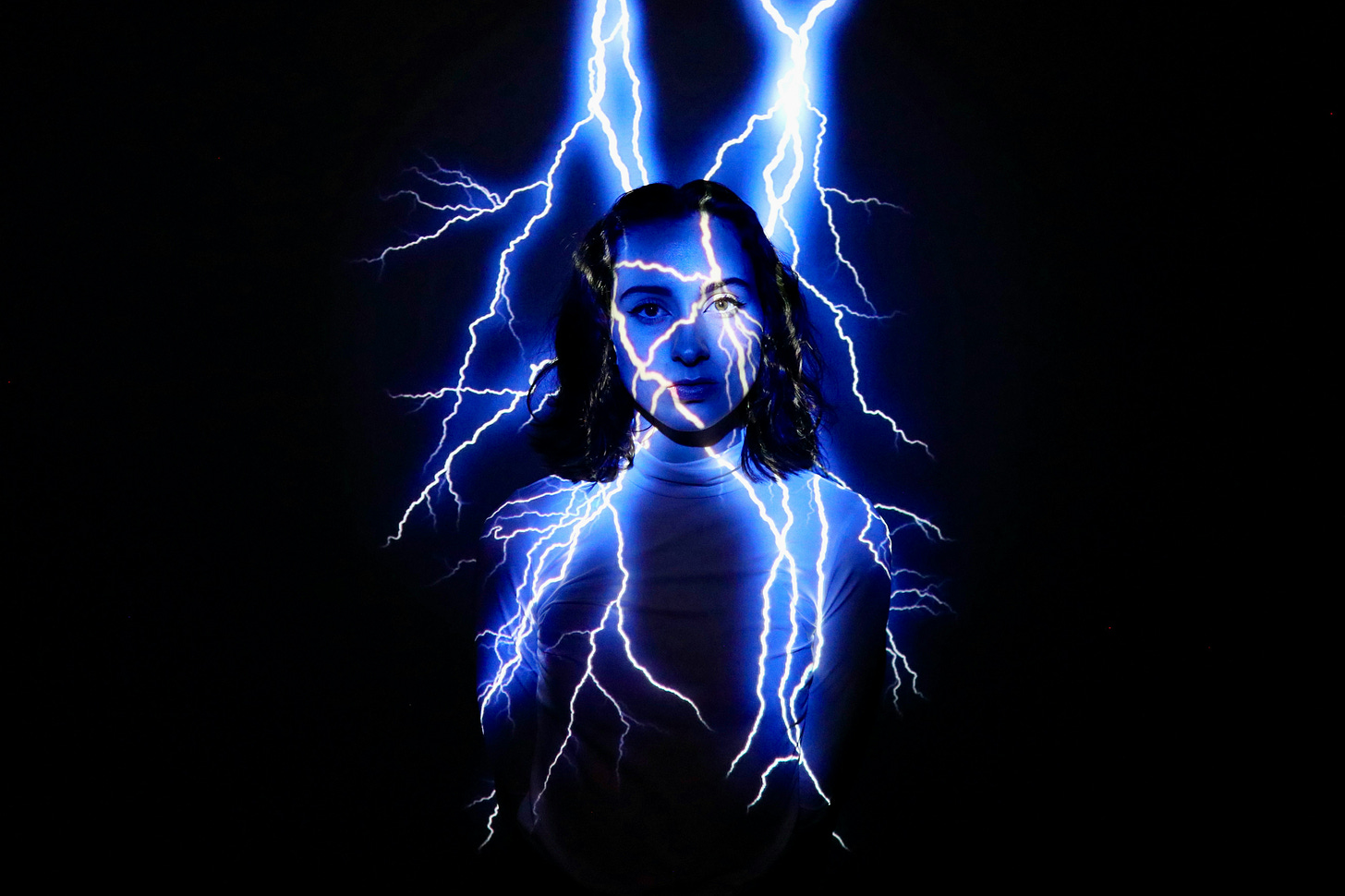 Brunette woman stands against a black background as white and blue bolts of lightning strike her head and spread through her body