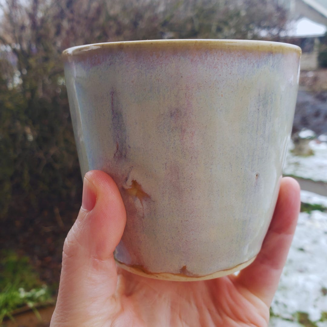 Close-up shot of Sarah's left hand, holding up the ceramic mug she is writing about. Out-of-focus plants and snow-covered grass can be seen in the background.