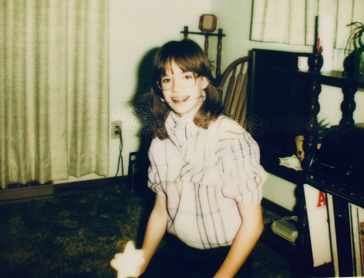 The author in big tortoiseshell glasses, braces and headgear in a pink-and-gray shirt with the collar flipped up. She sits on green shag carpet with a dark, knobby shelf behind her. If you look closely, you can see the A of the Annie album peeking out, first in line under the record player.