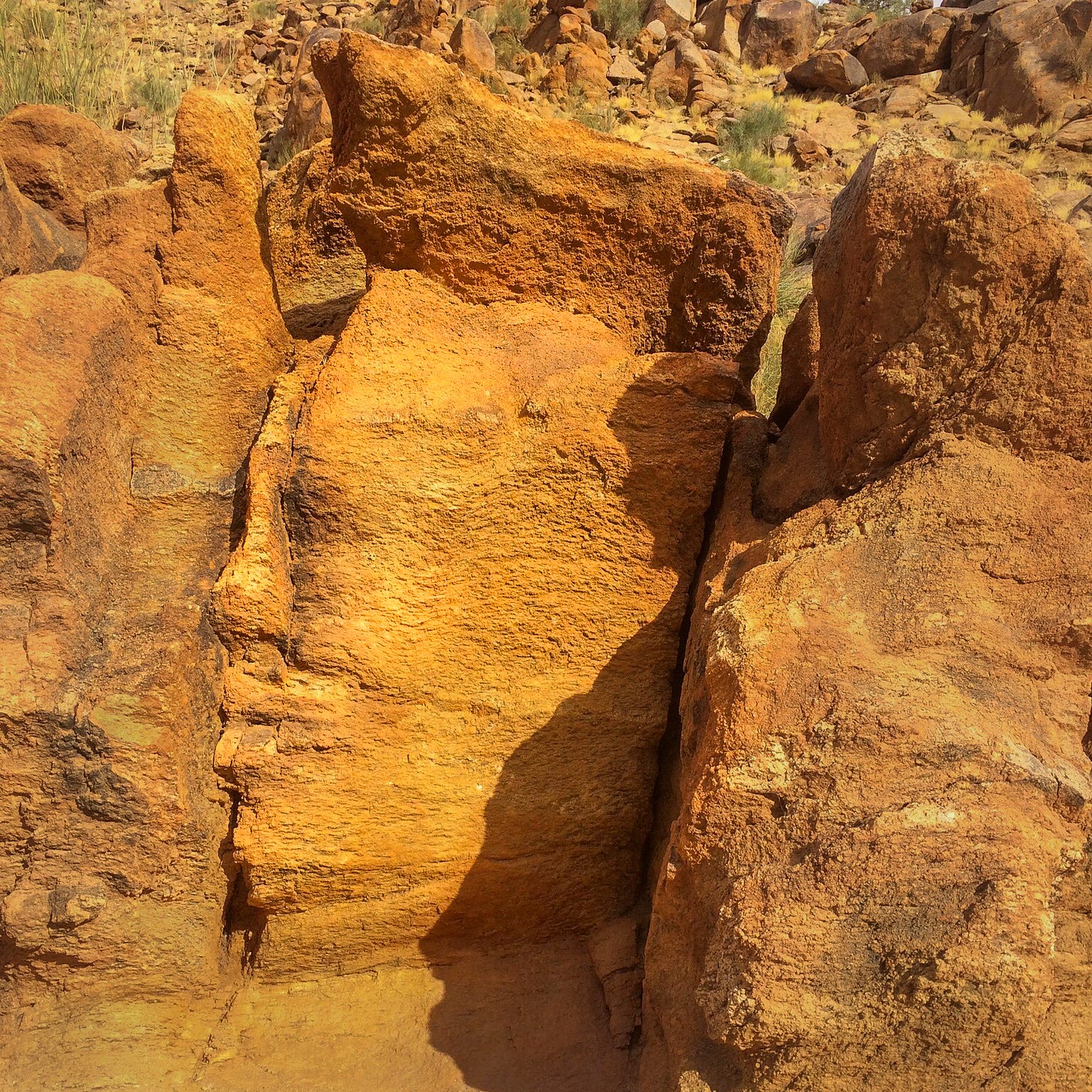 The photo shows a close-up of rugged, sunlit rock formations in a desert landscape. The rocks are predominantly orange and brown, with distinct layers and textures that suggest weathering over time. Shadows cast by the sun highlight the jagged surfaces, adding depth and contrast to the scene. Sparse tufts of dry grass can be seen in the background, suggesting an arid environment. The overall atmosphere of the image is warm, earthy, and ancient, evoking a sense of timelessness and natural resilience.