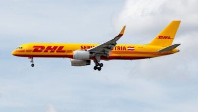 A mustard-yellow plane with red DHL lettering flies with wheels down.