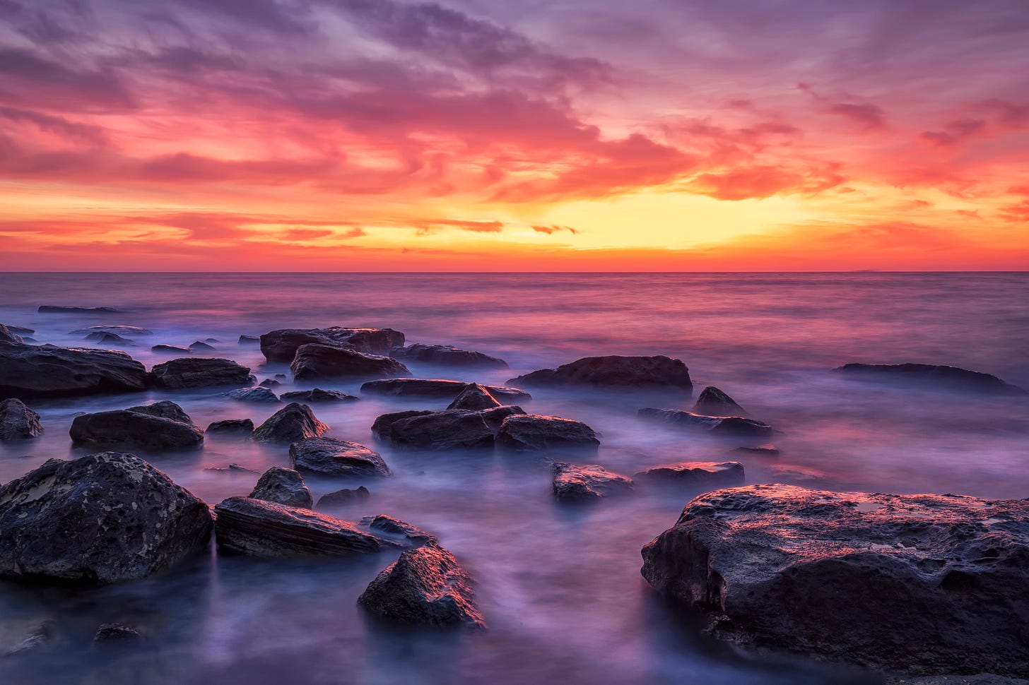 Stunning sunset over rocks on the sea shore, with red sky and purple sea