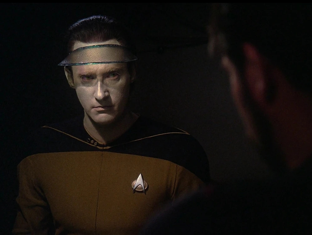 Data's poker face under a poker hat. He's looking at Riker who is out of focus in the foreground.