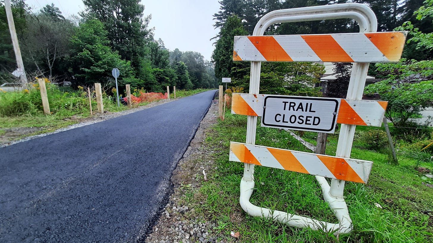 The Adirondack Rail Trail in Saranac Lake is seen with fresh blacktop laid on it, with a Trail Closed sign next to the fresh pavement. The trail is closed while under construction