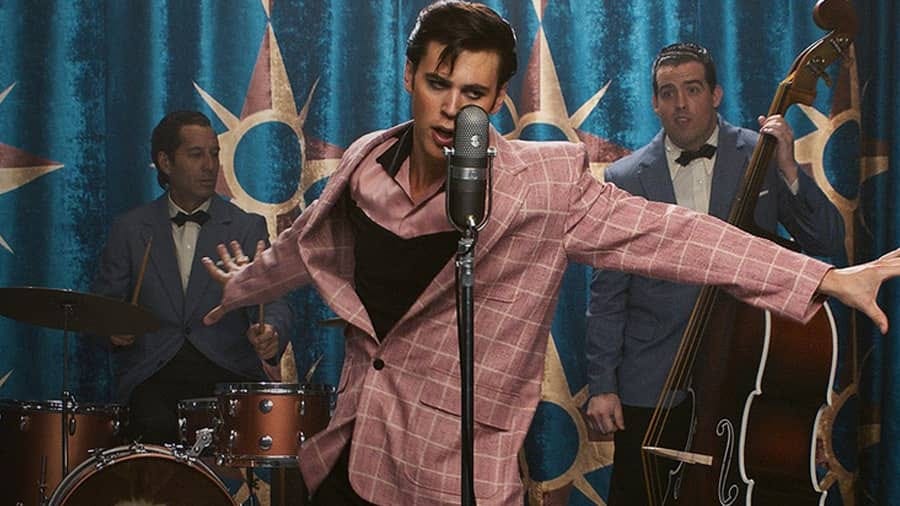 Still from Elvis showing Austin Butler in the titular role, wearing a pink checkered suit and performing on stage in front of his band.