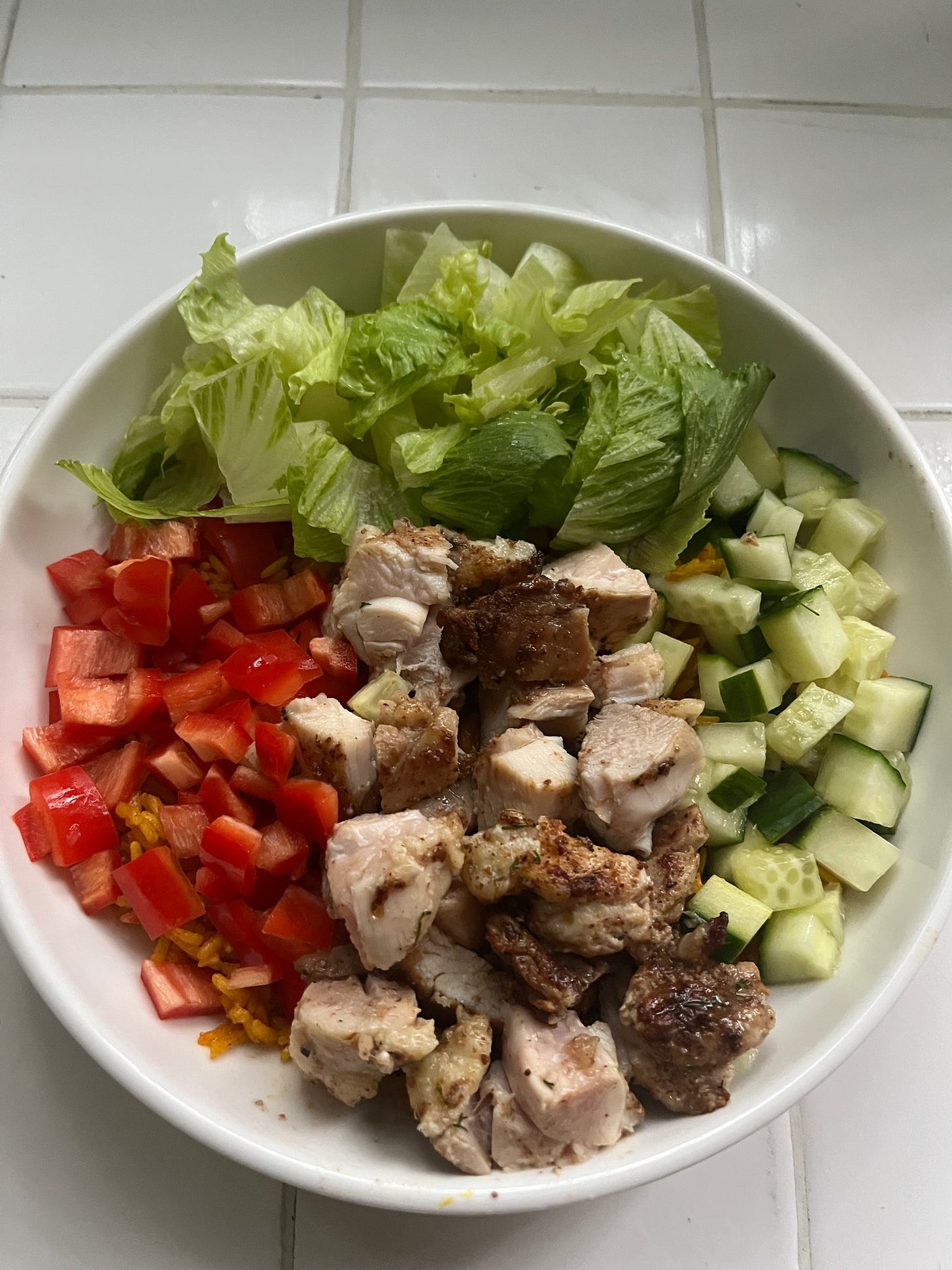 A bowl of yellow rice topped with seasoned chicken thighs, bell pepper, cucumber and lettuce
