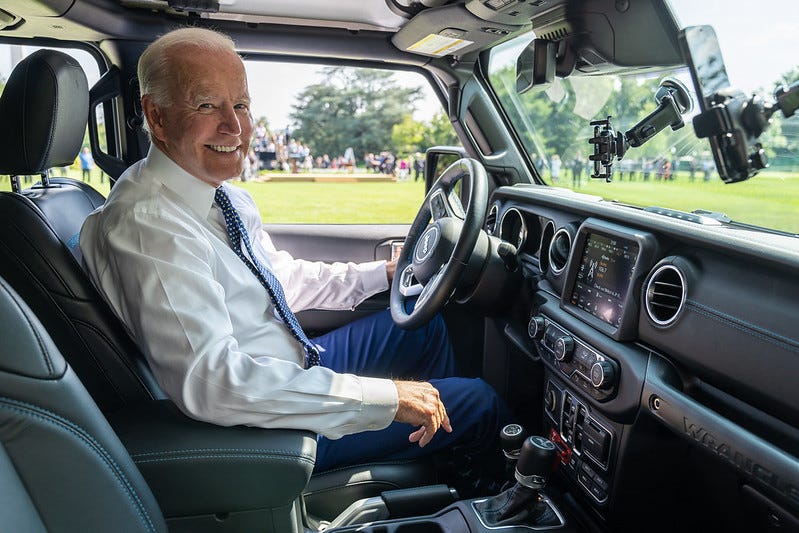 Joe Biden, in shirt and tie, smiles while sitting behind the wheel of an electric Jeep Wrangler at a White House EV event