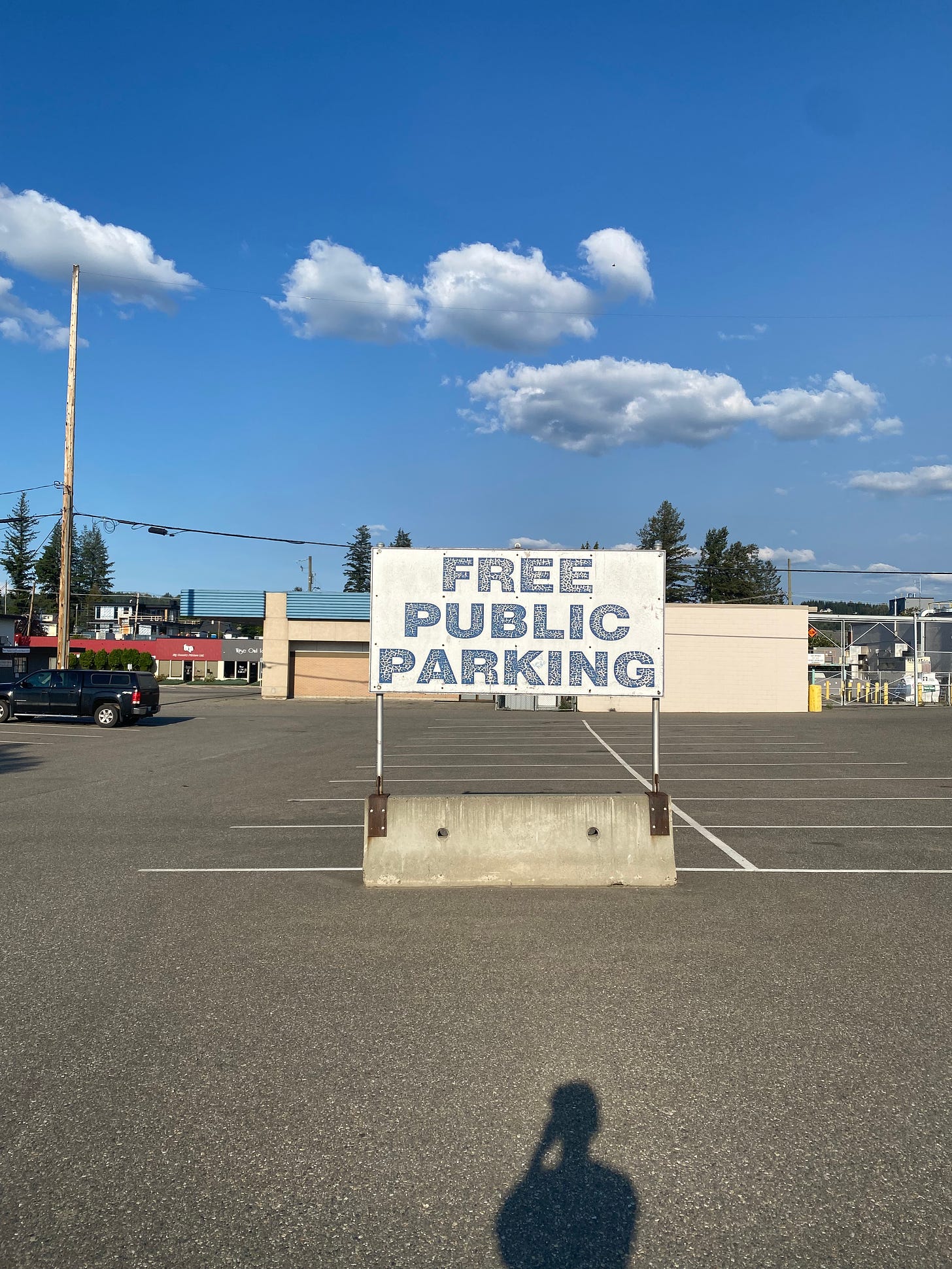 a nearly-empty parking lot with a faded blue and white sign in front that reads 'free public parking' in block letters. The sky is blue with a few clouds in the background.