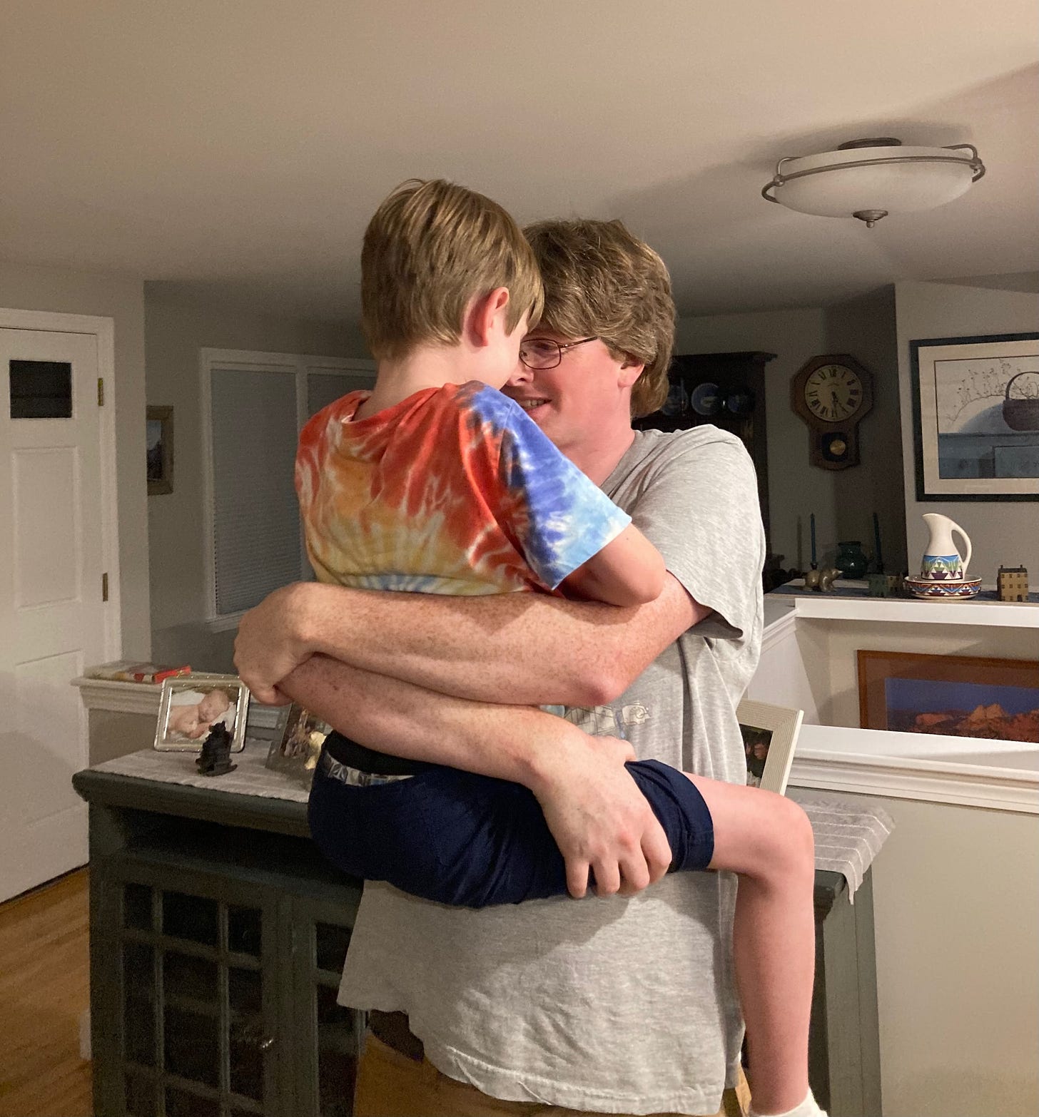 My husband (a white man in a white t-shirt) holding my younger son up (a white boy in a tie-dye shirt), standing in a living room