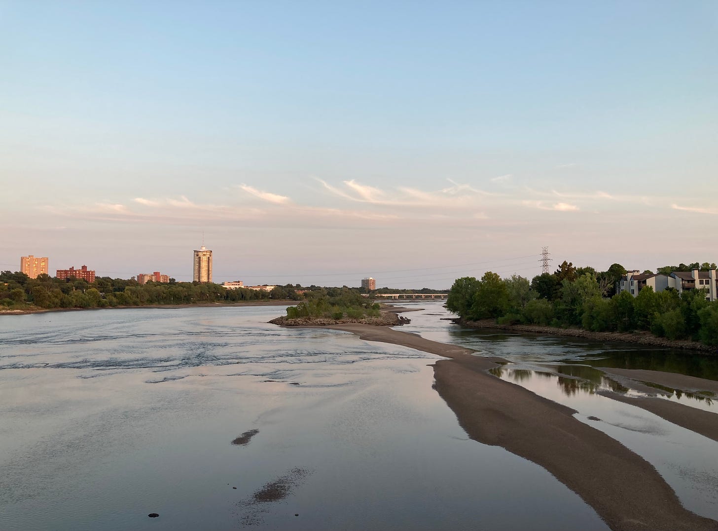 The photograph looks downstream over a wide, glassy Arkansas River, divided down one side by a bowing, terra cotta sandbar and vegetated island. The river reflects the fading pink and blue sky, streaked with delicate clouds just before dusk. In the distance, golden light illuminates the University Club Tower, Sophian Plaza and a few other buildings peeking out above the treetops along the east bank.  