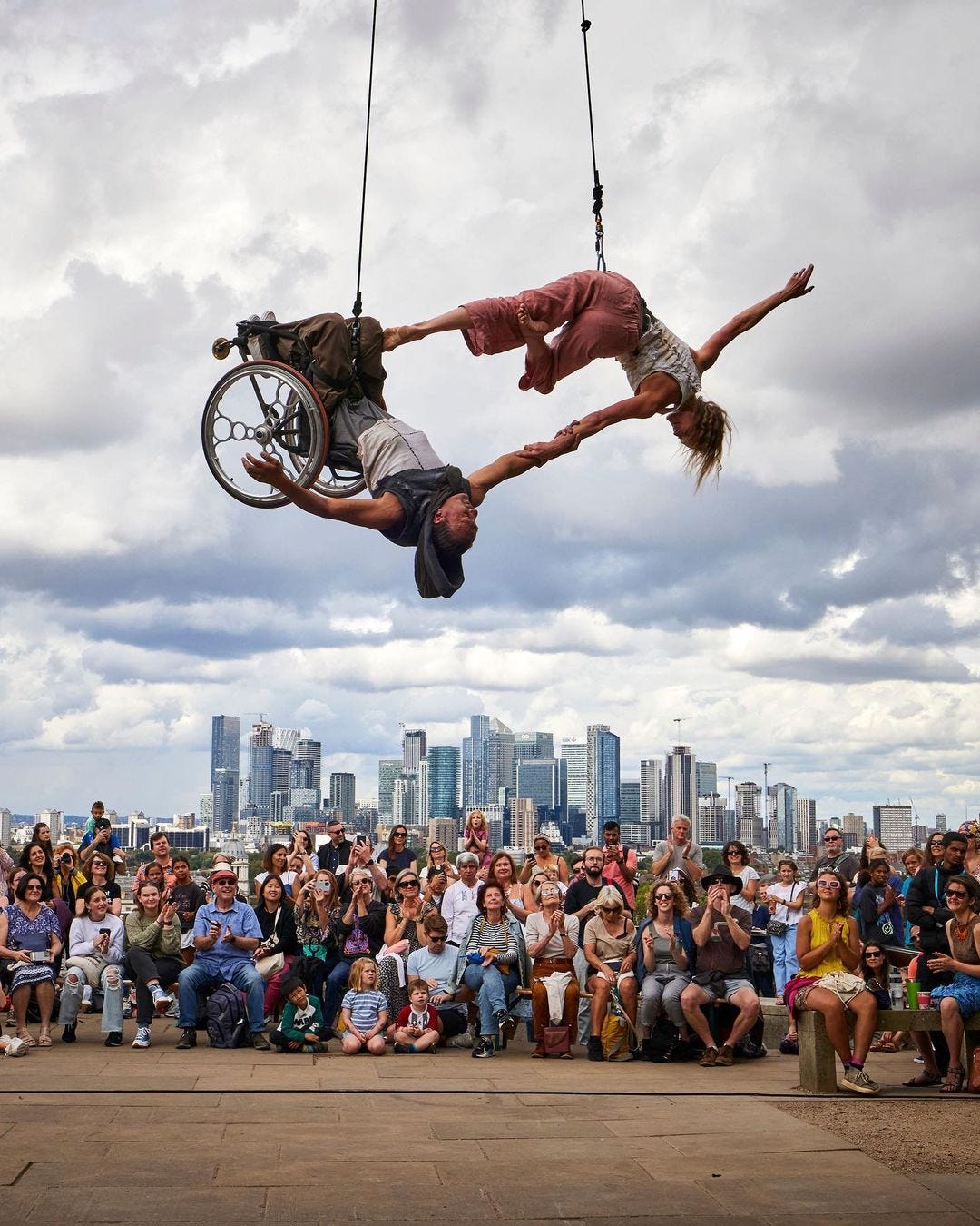 Two dancers, one in a manual wheelchair, suspend from cables that stretch out of the frame above. They are upside down over a hard stone surface with people on the ground looking up in awe, against a city skyline of buildings and dense, fluffy white clouds.
