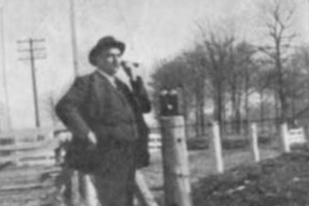 black and white photograph of a pan in a suit and bowler hat standing in front of a barbed wire fence talking on the phone