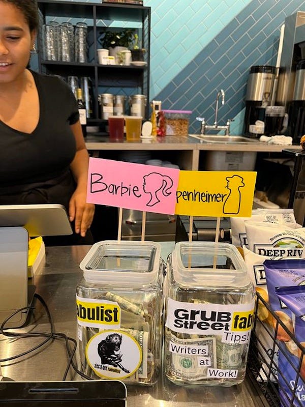 Photo taken in a coffee shop. We see a barista ringing up a sale. Before her, on the counter, are two jars for tips, each with a hand-lettered sign. On the left, a pink sign has the silohuette of a pony-tailed woman and the word, "Barbie." On the right, the jar has a yellow sign, the silohuette of a man wearing a fedora & smoking a cigarette, with the word Oppenheimer" written on it. 
