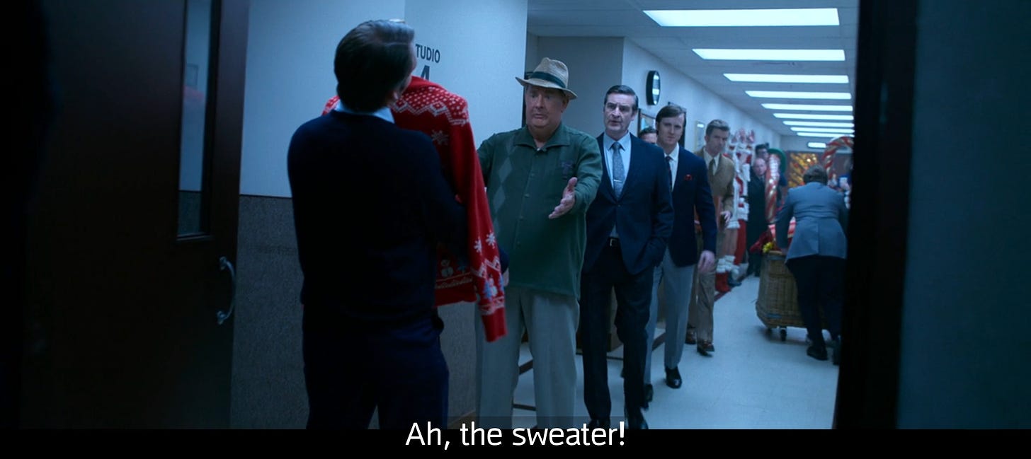 Tom Hanks as Colonel Tom Parker (center) gesturing toward a red Christmas sweater held by a network exec while TV employees run around behind him, in ELVIS. Dialogue from Parker captioned on the image: Ah, the sweater!