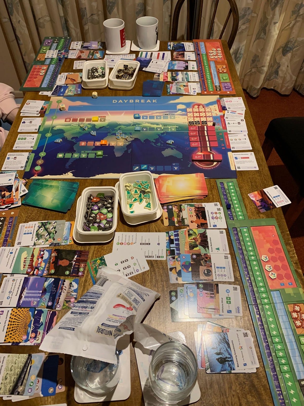 A photo of a dining table with the Daybreak board game laid out. There are many colourful tokens, cards, and world map, and a big thermometer - plus a bag of lollies.