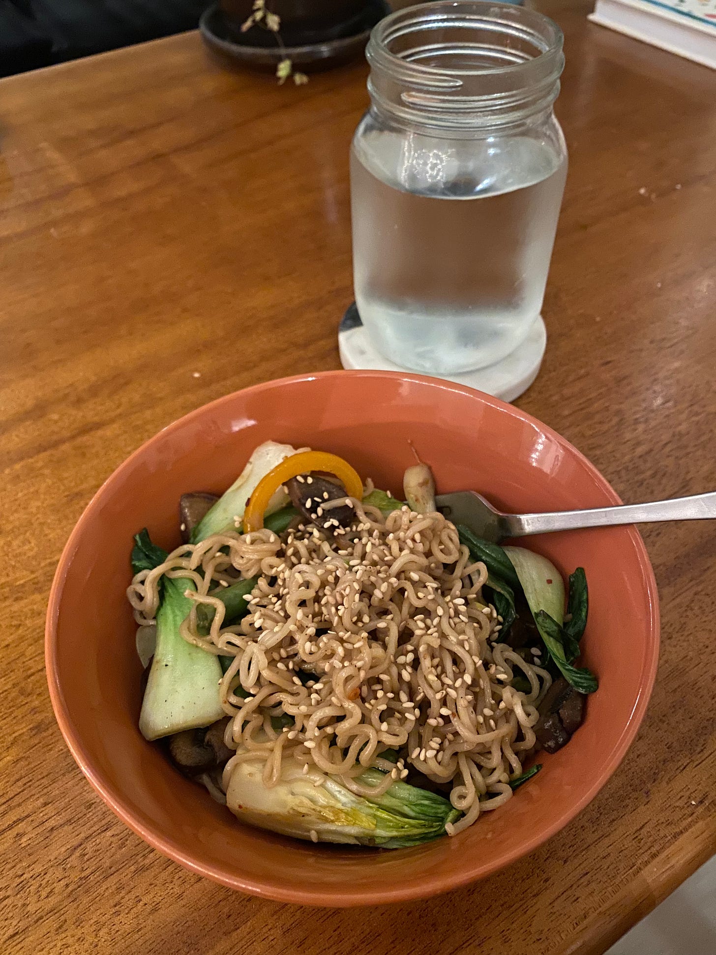 The stir-fry described above in an orange bowl with a fork sticking out of it: thin ramen noodles, quartered pieces of bok choy, slices of pepper and mushrooms, pieces of green beans, all dusted with sesame seeds.