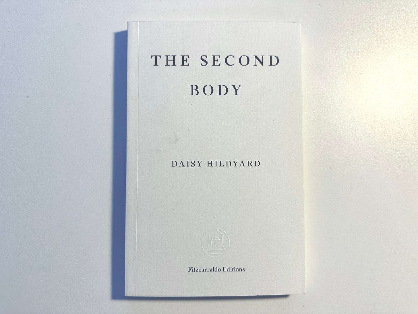 Picture of a book titled "The Second Body" by Daisy Hildyard, on a white table. The cover is plain white with no images with the title and written in capital serif text.