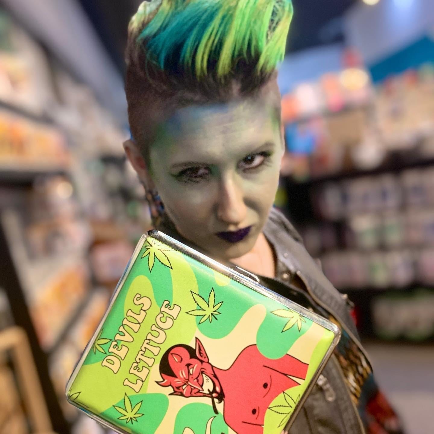 Photo of Lyric, in a store, dressed in green, with short spiked green hair, holding up a card that says “the Devil’s lettuce” while making a serious face 