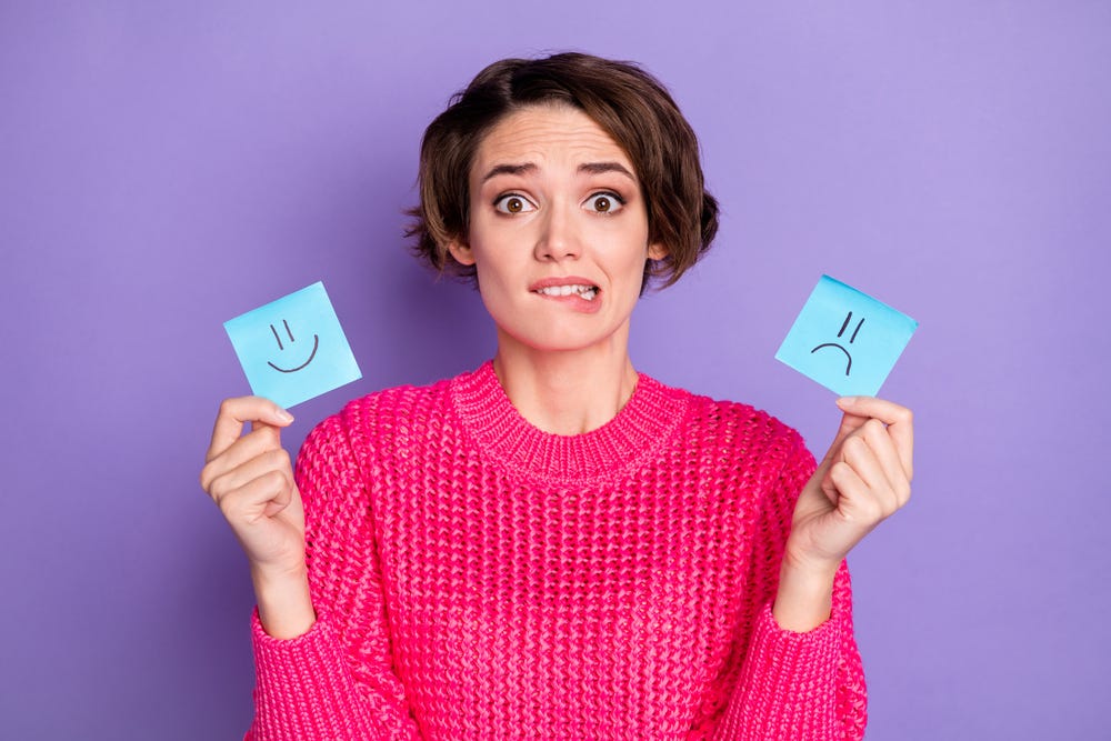 Woman with short dark hair wearing a hot pink sweater, making a confused face and holding up two post-it notes, one with a happy face on it and one with a sad face on it.