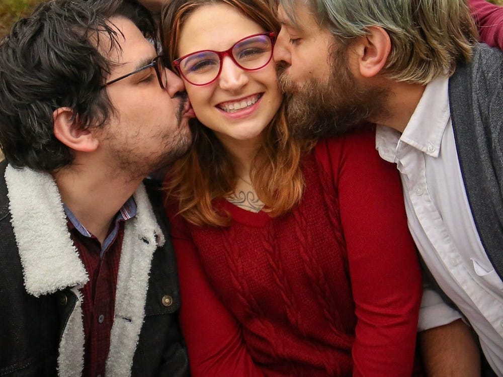 ohhh this is a photo of yours truly wearing a red sweater and being kissed by my partner Ty on the left check (well, my right, your left) while my husband Daniel is kissing me on the right (my left) cheek. I'm cheesing big time bc I love them so much